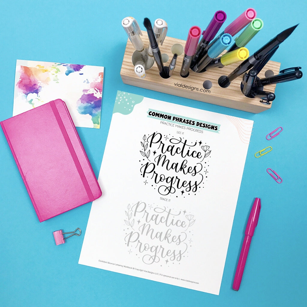 Stationery items, including a pink notebook, colorful markers, and a sheet of calligraphy practice paper on a blue background. Practice makes progress bounce lettering design practice page